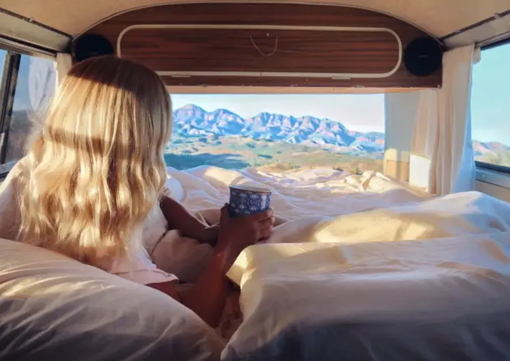 Lady laying down in a campervan bed with a mug in hand, and mountain ranges in the distance. 
